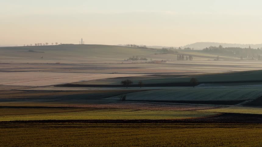 A wide rural landscape of rolling agricultural fields stretches across gentle hills under soft dusk light