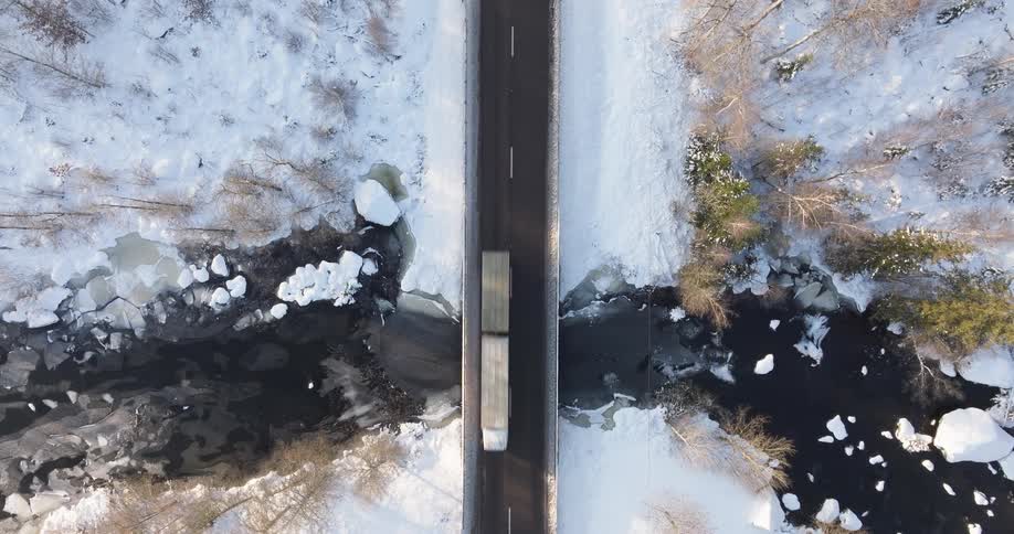 An aerial view shows a large truck as it drives across a bridge over a river with chunks of ice while surrounded by a forest covered in white snow during the day
