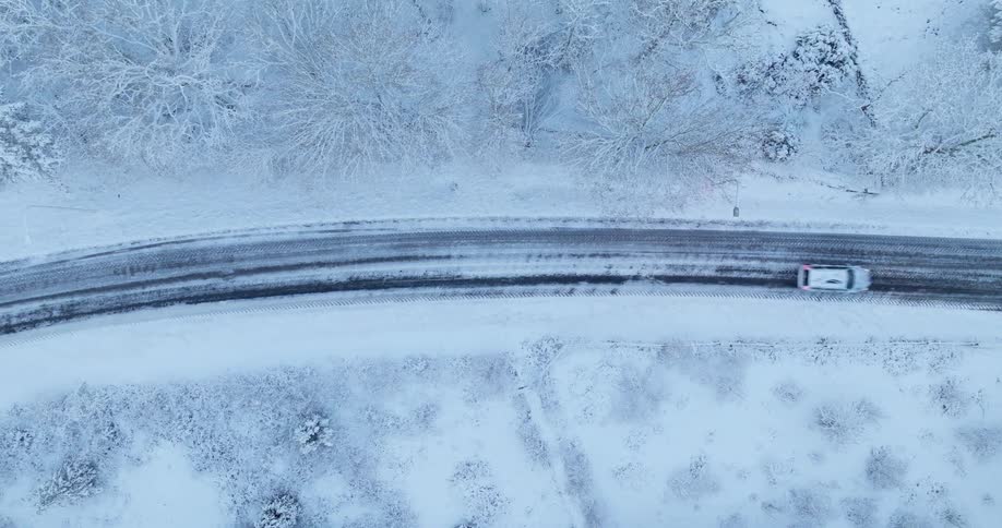 This aerial shot shows a single vehicle traveling along a dark slushy road that cuts through a pristine snowcovered landscape the contrast between the black asphalt and the surrounding white trees creates a quiet wintery atmosphere