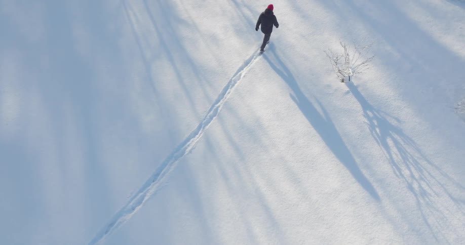 An aerial view shows a person wearing a red hat as they walk across a field covered in white snow while leaving a trail of footprints during a sunny day