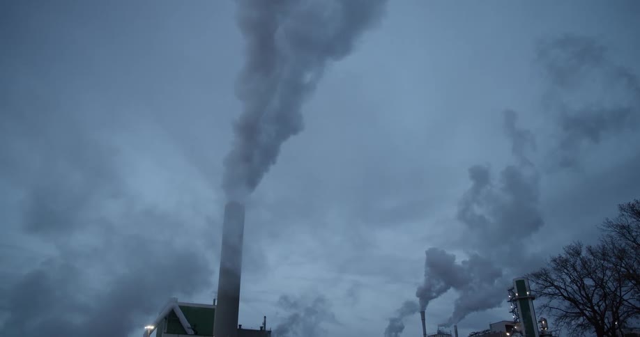Thick plumes of smoke rise from factory chimneys into a cold overcast sky casting an industrial landscape in a bleak heavy mood