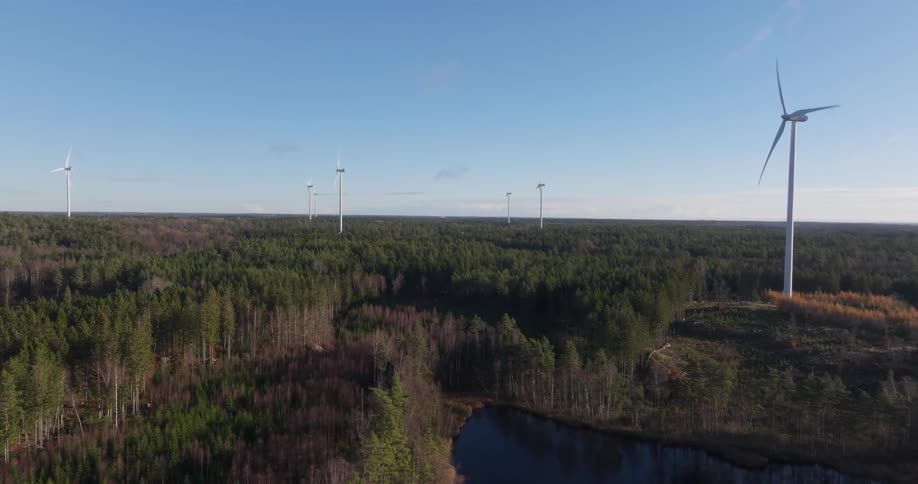 An aerial view shows several white wind turbines spinning above a dense green forest with a small blue lake during a clear day