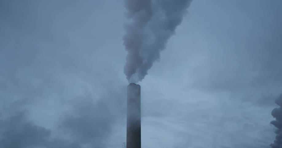 A view shows a tall chimney as thick gray smoke rises from it into a cloudy sky during the day