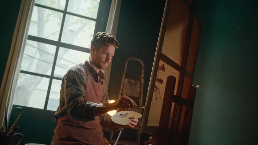 An artist in a brown apron sits in a sunlit studio carefully applying paint to a canvas on a wooden easel the scene captures a moment of quiet focus as he mixes colors on his palette while natural light streams through a large window