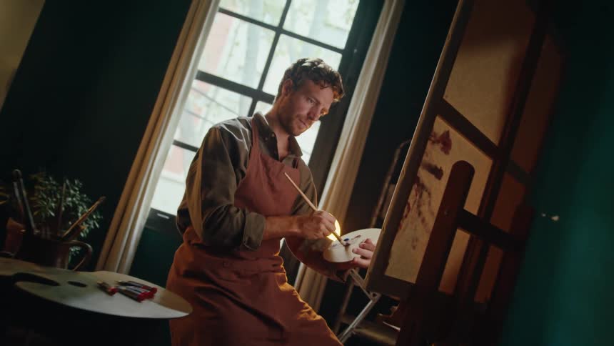 Man mixes paint on a palette while working in front of an easel in a studio