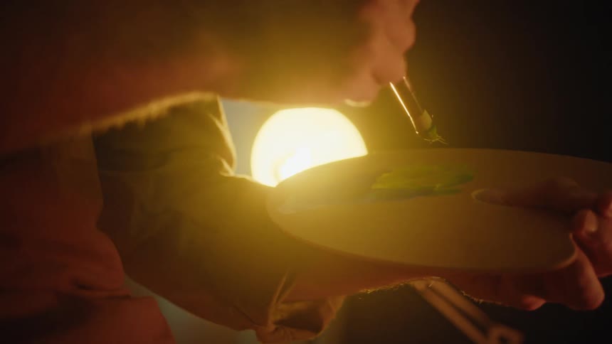 Brush mixes green paint on a palette held by a hand under intense warm lighting
