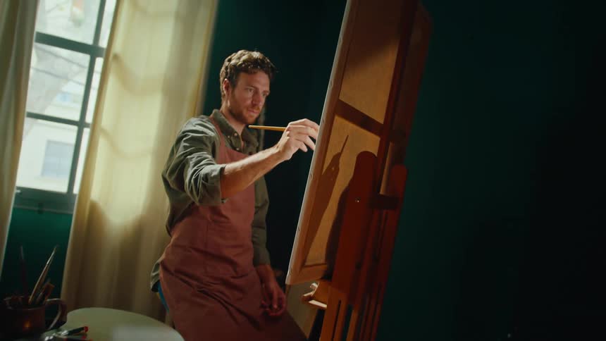 Man paints on an easel inside a studio with artificial lighting and art materials around