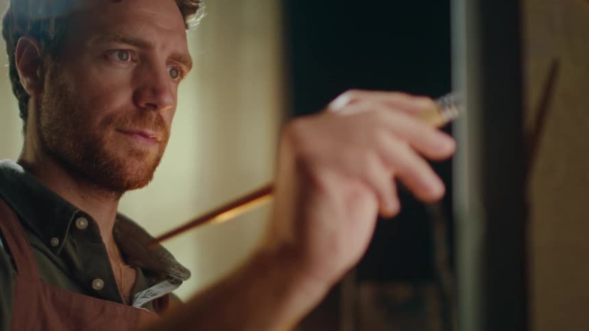 A close view shows a man with brown hair and a beard focused on painting with a brush in an art studio
