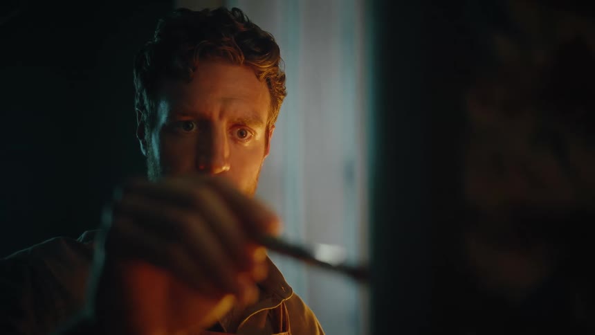 Man holds a brush in front of his face while working on an outoffocus surface under warm lighting