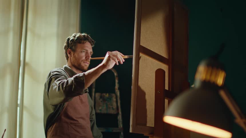 Man applies paint with a brush to a canvas mounted on an easel inside a studio