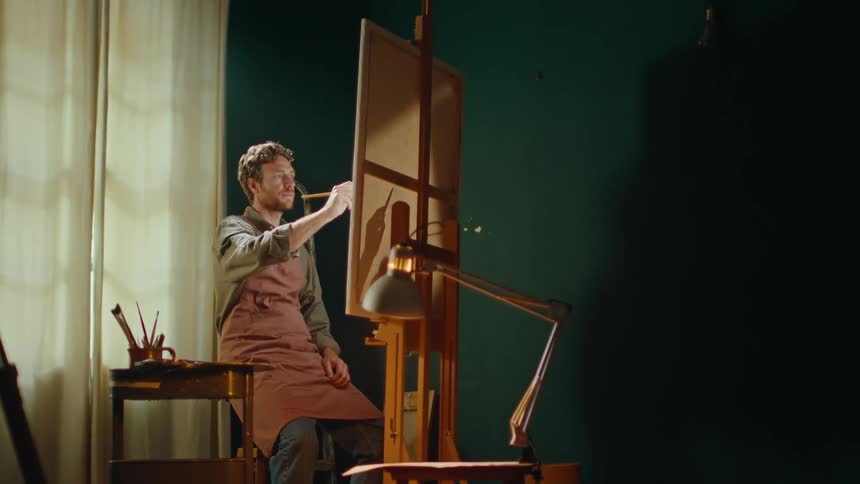 The man looks over the canvass then sits to paint