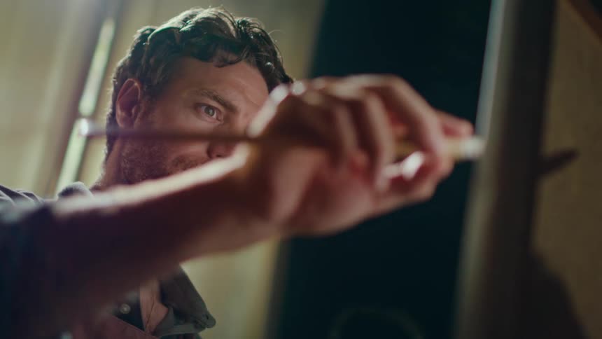 An artist with wavy hair and a beard works intently on a painting he uses a paintbrush to apply careful strokes to a canvas while bathed in warm light