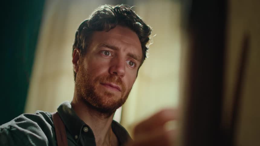 A man with wavy hair and a beard focuses intently on his work while painting on a canvas he uses a paintbrush to apply careful strokes with warm light highlighting his concentrated expression