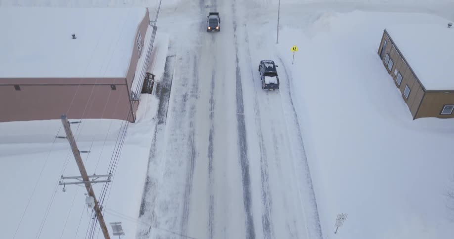 Cars on the side of a snow covered road with light traffic in the neighborhood along the coast and mountains in the distance