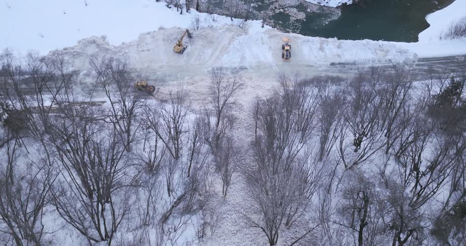 Heavy machinery clears snow from a frozen roadway near a body of water in a winter landscape seen from above