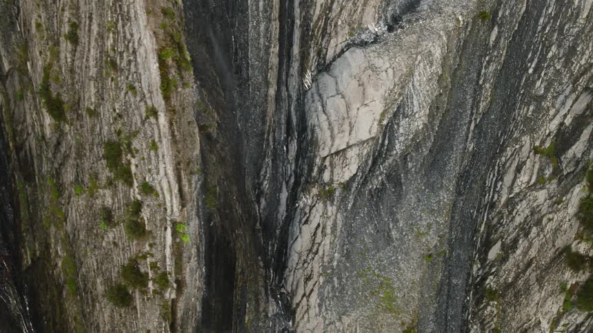 An aerial top down view captures the rugged textures of a steep gray cliff face where vertical ridges and rocky crevices create a dramatic natural pattern interspersed with small patches of green vegetation and loose mountain debris