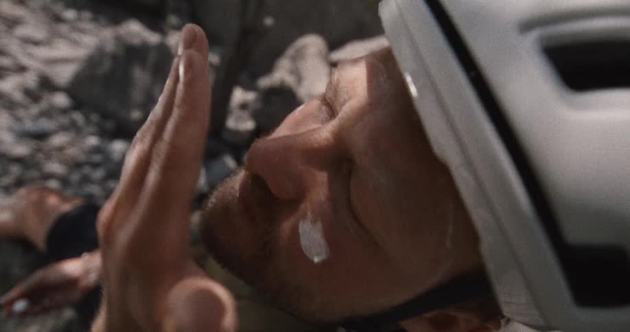 A close up view shows a person wearing a white helmet as they apply white cream to their face while sitting on a rocky area during a sunny day