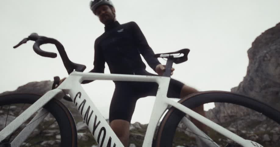 A view shows a person wearing a helmet and a black long sleeve shirt as they mount a white bicycle on a rocky mountain during the day