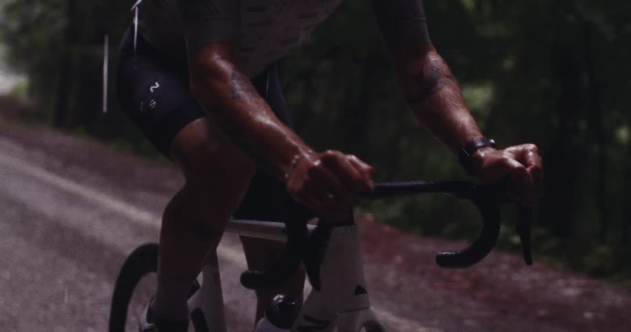 A cyclist with tattooed arms grips the handlebars while riding along a dark wet road