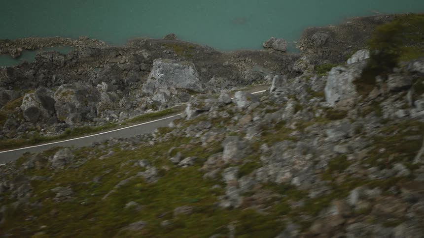 An aerial camera follows a lone cyclist as he rides along a narrow paved road carved into a steep mountain ridge overlooking a turquoise glacial lake and rocky terrain