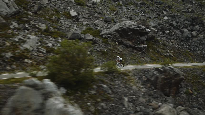 A cyclist wearing a white helmet and professional gear rides a road bike quickly along a narrow paved highway that cuts through a vast mountain landscape filled with large gray boulders and sparse green vegetation on a cloudy day