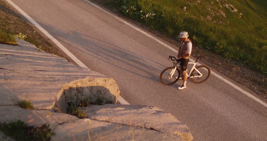 Cyclist stands beside a bicycle on a mountain road lit by golden lateafternoon light
