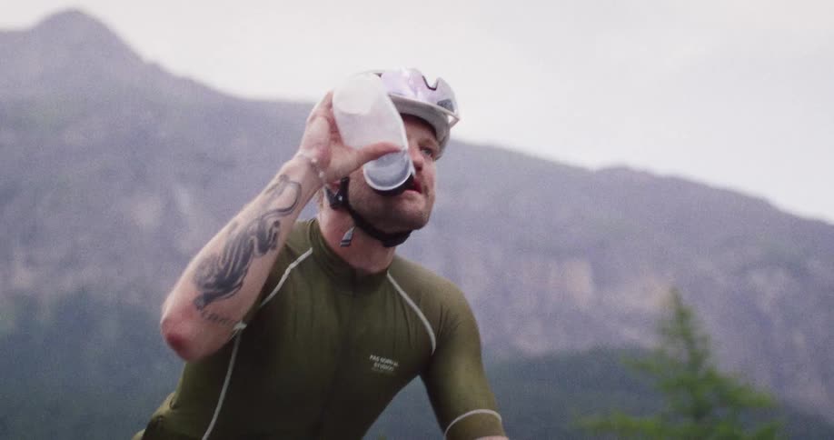 A male cyclist in a green jersey rides a white bicycle up a mountain road takes a plastic bottle from the frame cage drinks water while pedaling and then looks ahead toward the cloudy horizon