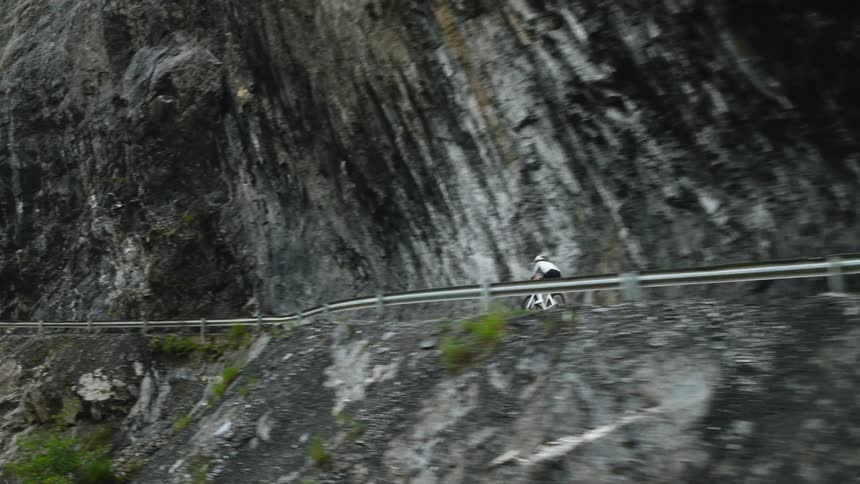 A view shows a person wearing a white helmet as they ride a bicycle along a road carved into a dark rocky cliffside during the day
