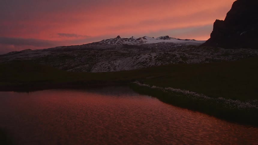 A vibrant red sunset reflects off a winding mountain stream leading toward snow capped peaks