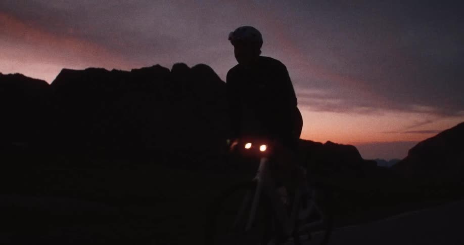Silhouetted cyclist with front lights on rides along a mountainous road at dusk