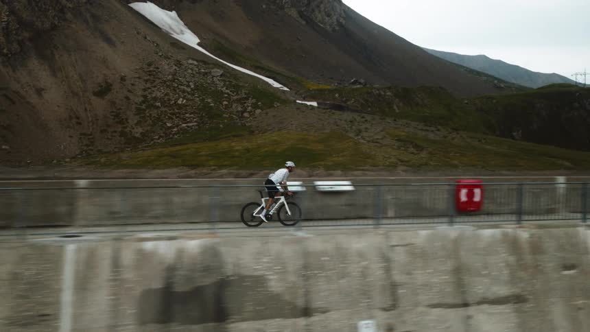 A view shows a person wearing a helmet and a white jersey as they ride a bicycle along a concrete wall next to a large body of water and a rocky mountain during the day
