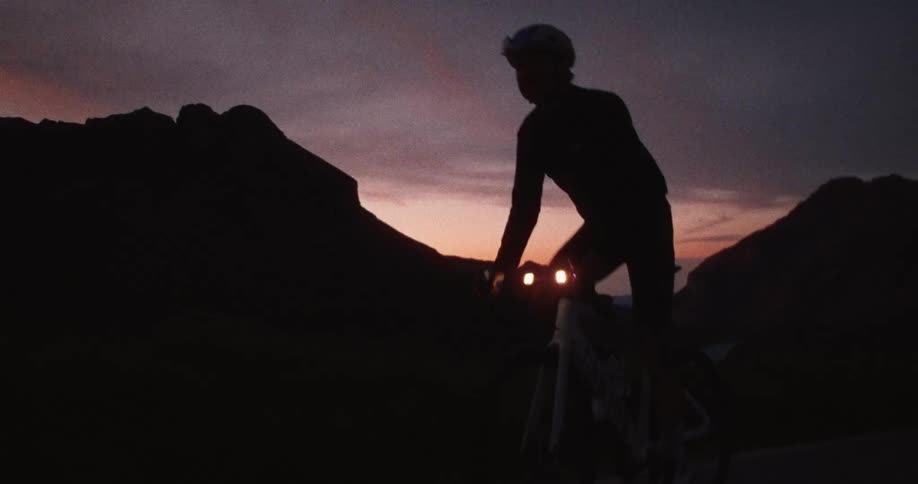 A cyclist rides a road bike along a winding mountain pass during a dark orange sunset as the silhouette of jagged peaks stands against the evening sky while front lights illuminate the path ahead during the descent