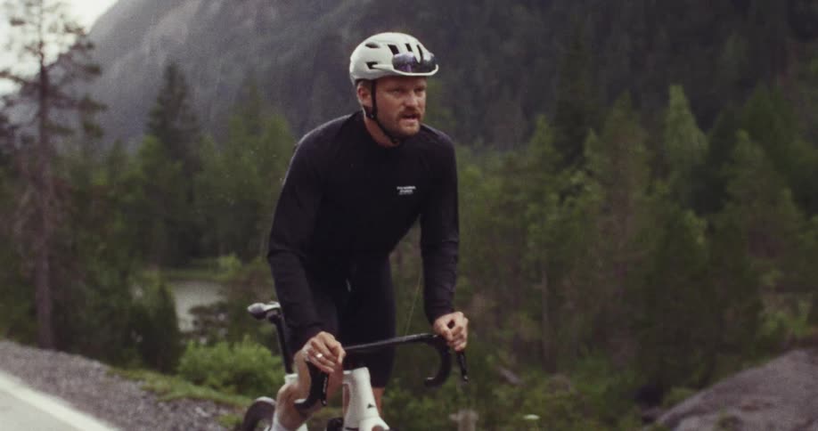A view shows a person wearing a helmet and a black long sleeve shirt as they ride a bicycle along a road during a rainstorm in a forest