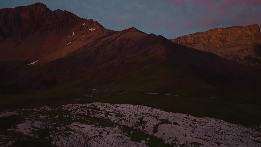 A view of a mountain range at dawn with pink clouds in the sky above dark rocky slopes and a foreground of light colored stones