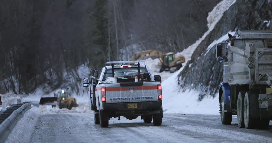 A maintenance truck drives along a snowy mountain road and parks so a man can exit while construction vehicles work to clear debris and stabilize the icy roadside