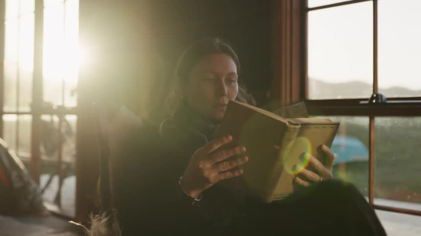Woman seated indoors reading a book near a window with natural light in the background