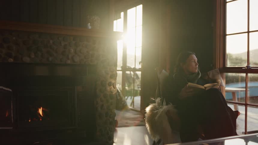 Person seated indoors beside a lit fireplace holding a book with natural light coming through the window