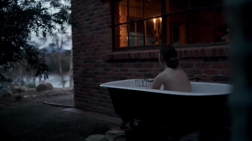 A woman sits in an outdoor clawfoot bathtub next to a brick house at dusk