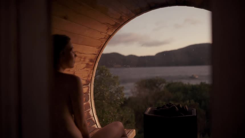 Woman seated inside a wooden interior looking at the outdoor landscape through a circular opening at dusk