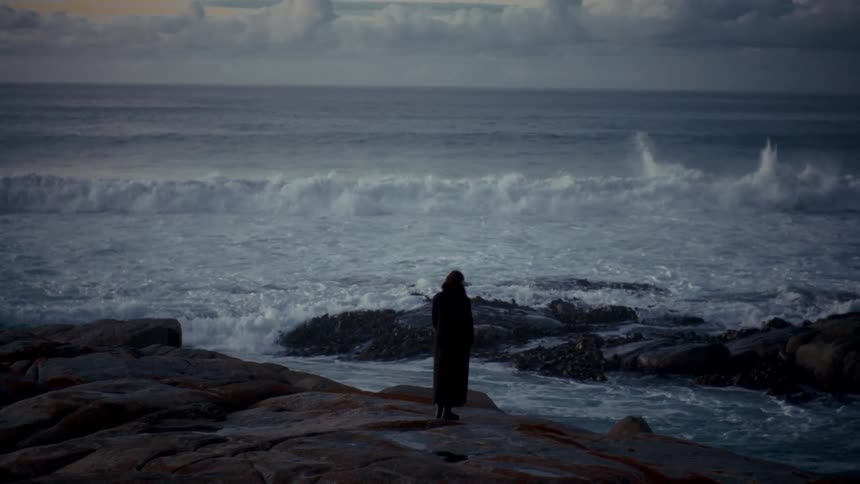 Woman stands along the coast as splashing waves roll in to shore and crash against rocks