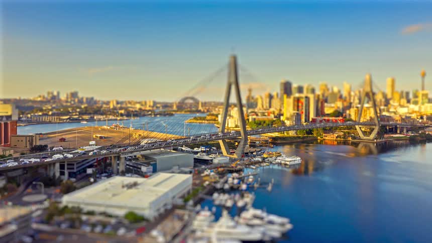Aerial view of a modern cable stayed bridge spanning a harbor with a sunlit city skyline in the background