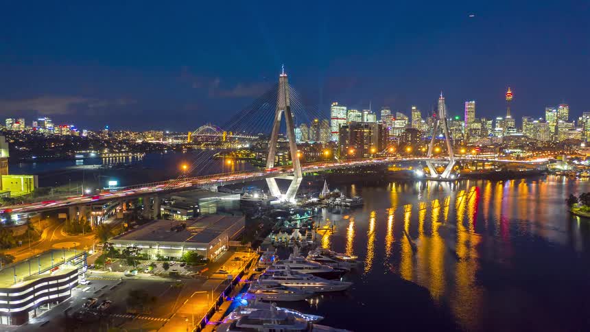 Bridge with heavy traffic over a river with a marina to the side and a metropolitan city on the background outdoors during dusk and night time