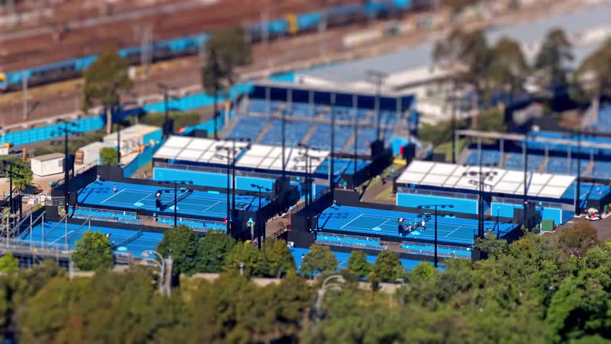 A highangle tiltshift effect transforms the scene into a miniature world capturing tiny figures rallying on vibrant blue tennis courts while a train speeds along the railway tracks in the background