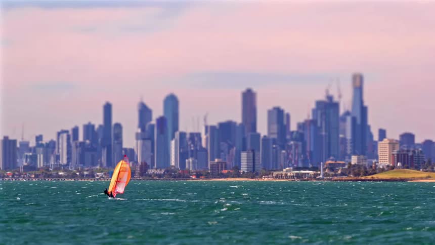 A lone windsurfer glides across choppy blue water with a modern city skyline stretching across the horizon behind them