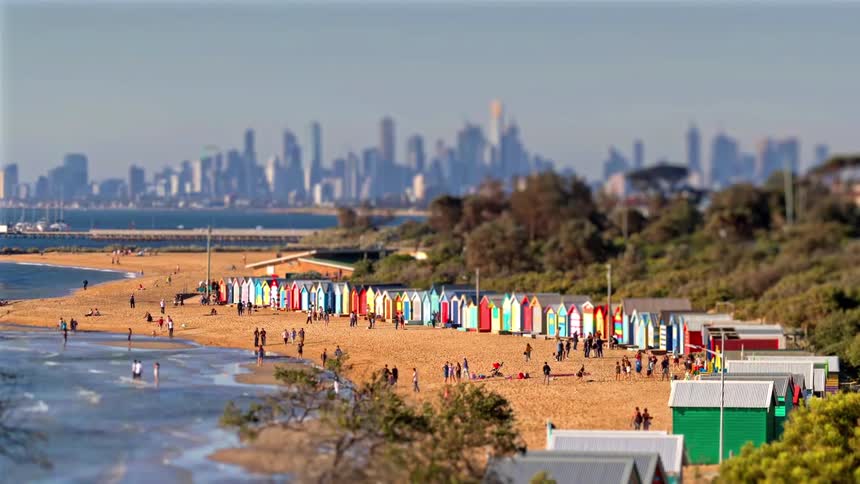 A view from a distance shows many small beach huts painted in different colors on a beach as people walk around them near a large body of water and a city with many tall buildings during the day