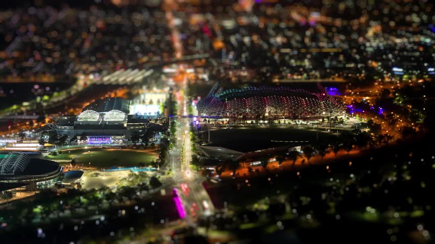 A nighttime aerial view captures a brightly lit stadium complex and surrounding city streets glowing with colorful lights and traffic