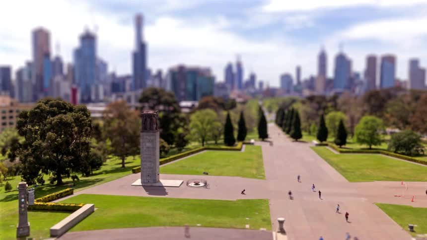 Landscaped axis with a central monument paved walkways and people moving through the area with an urban skyline in the background