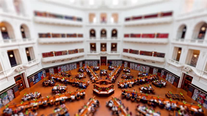 A view from above shows many people as they sit at desks and read books in a large library with several floors during the day