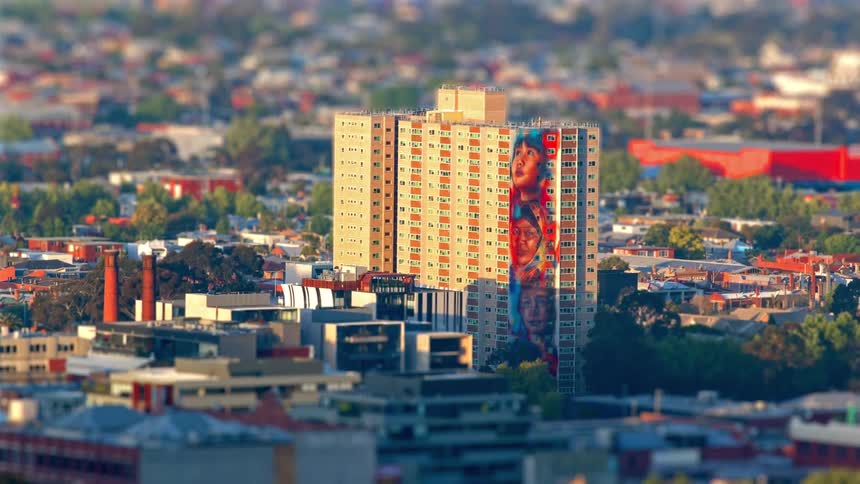 A tall apartment building with a striking mural of faces rises above a densely packed neighborhood standing out against the softly blurred cityscape around it