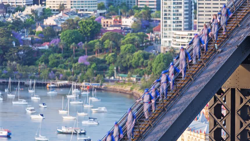 A group of people wearing the same blue jackets walking single file along a bridge walkway with several sailboats docked in the water below at midday